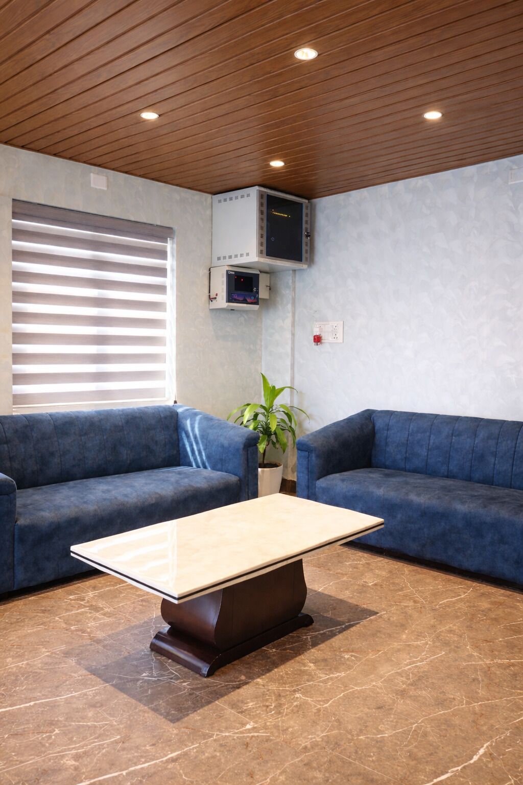 A modern lobby with two blue sofas, a marble-tiled floor, a wooden ceiling, and a small potted plant.