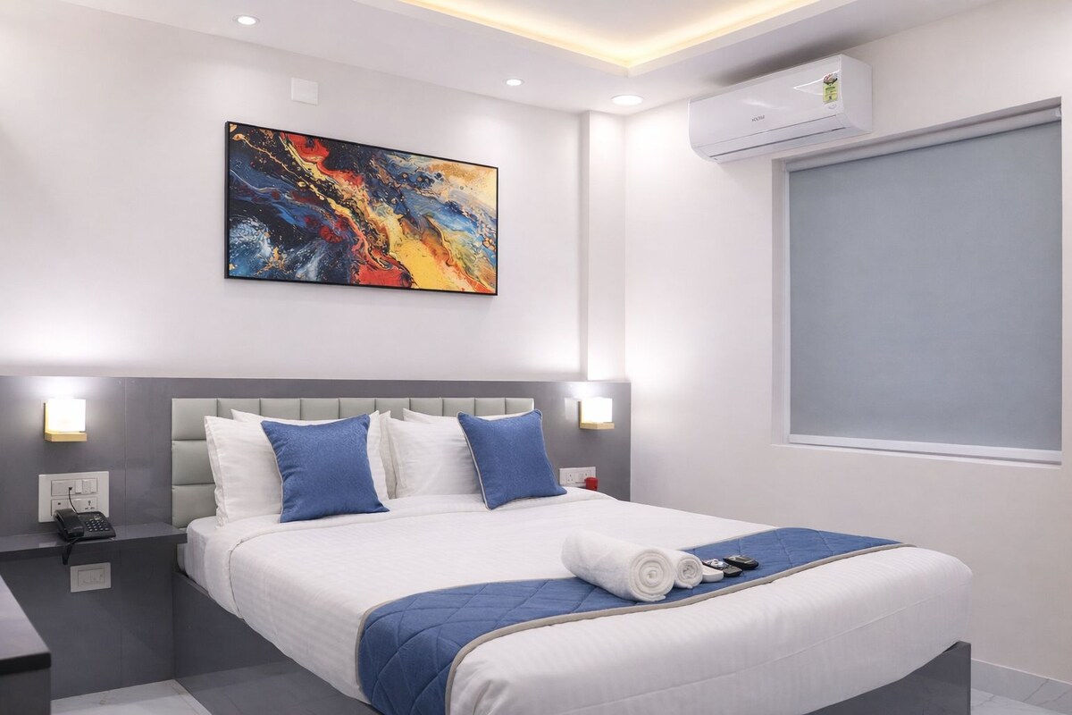 A modern hotel room with a large bed, a colorful abstract painting, a wall-mounted air conditioner, and a window with a view.