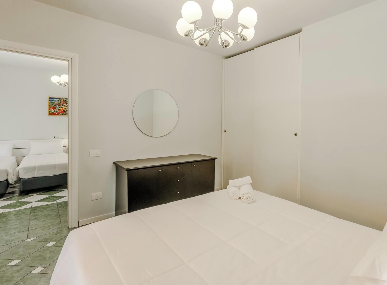 Quarto de hotel com cama, penteadeira, espelho redondo e porta que dá para outro quarto.