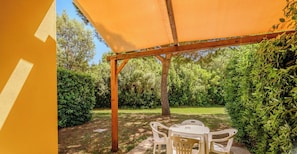 Une terrasse couverte, agrémentée d’une table et de chaises, entourée de verdure et d’arbres.