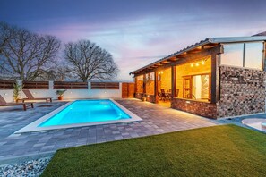 Un espace piscine extérieur moderne, doté d’un patio en pierre, d’un coin salon abrité et d’une pelouse bien entretenue.