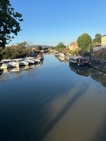 A calm river with boats docked on the left, a bridge in the distance, and buildings along the riverbank.