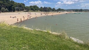 Ein Strand mit badenden und sonnenden Personen, eine Rasenfläche im Vordergrund und ein klarer Himmel mit Wolken.