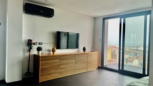 Een moderne woonkamer met een grote flatscreen televisie aan de muur, een houten console met decoratieve objecten, en een schuifdeur naar een balkon met uitzicht op de stad.