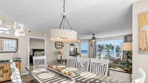 Uma sala de jantar com uma mesa comprida, cadeiras brancas e um lustre.