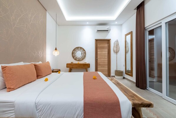 A modern bedroom with a large bed, a wooden bench, a small table with a mirror, and a sliding glass door leading to a balcony. - Villa Terracotta (Tabanan)