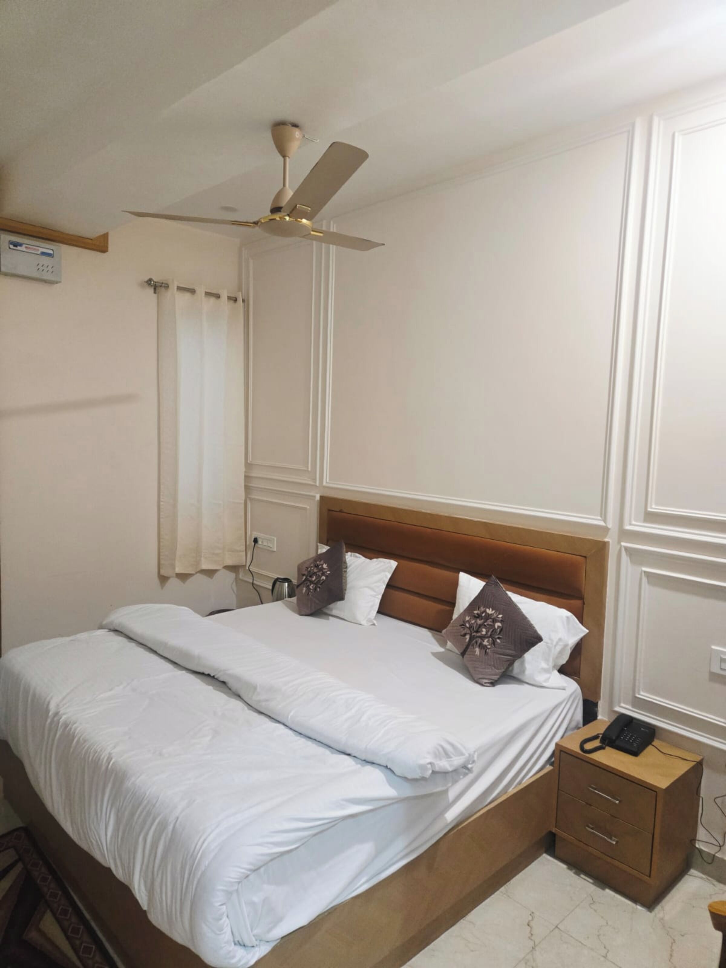 A neatly made bed with white linens, a wooden headboard, and decorative pillows. There is a bedside table with a phone and a small drawer. A ceiling fan is visible above the bed.
