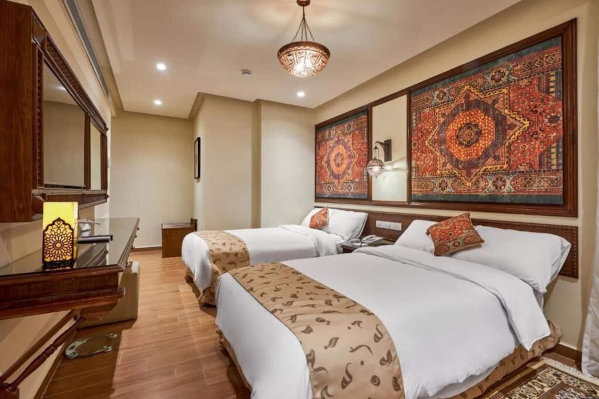 A hotel room with two beds, a wooden desk, a mirror, and a large decorative tapestry on the wall.