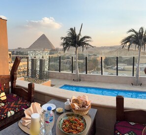 A balcony with a pool, a table with food and drinks, and a view of the pyramid. - 7 Gates Boutique Hotel Pyramids (Giza)