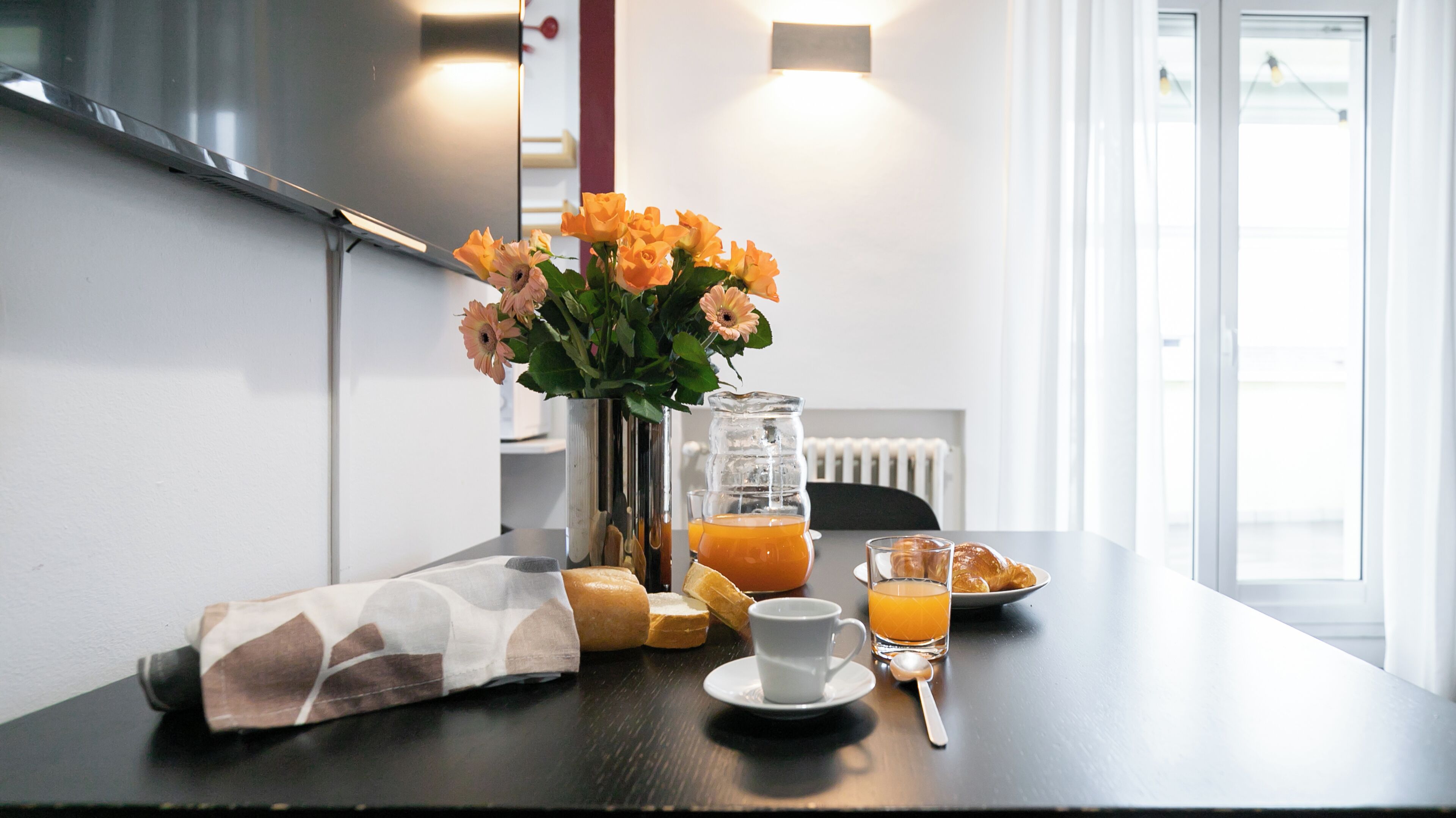 A breakfast setup with a vase of flowers, a glass pitcher of orange juice, a cup, a plate with croissants, and a napkin on a table.
