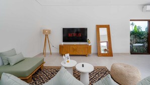 Moderni olohuone, jossa on vihreä sohva, puinen TV-taso, suuri litteänäyttöinen televisio, jossa näkyy teksti ’NETFLIX’, pieni pyöreä pöytä ja suuri peili.