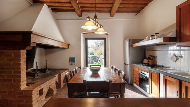 A rustic kitchen with a brick fireplace, wooden dining table, and a view through the door. - Cipressina – Spacious Tuscan Apartment with Pool for Families and Groups (Montespertoli)