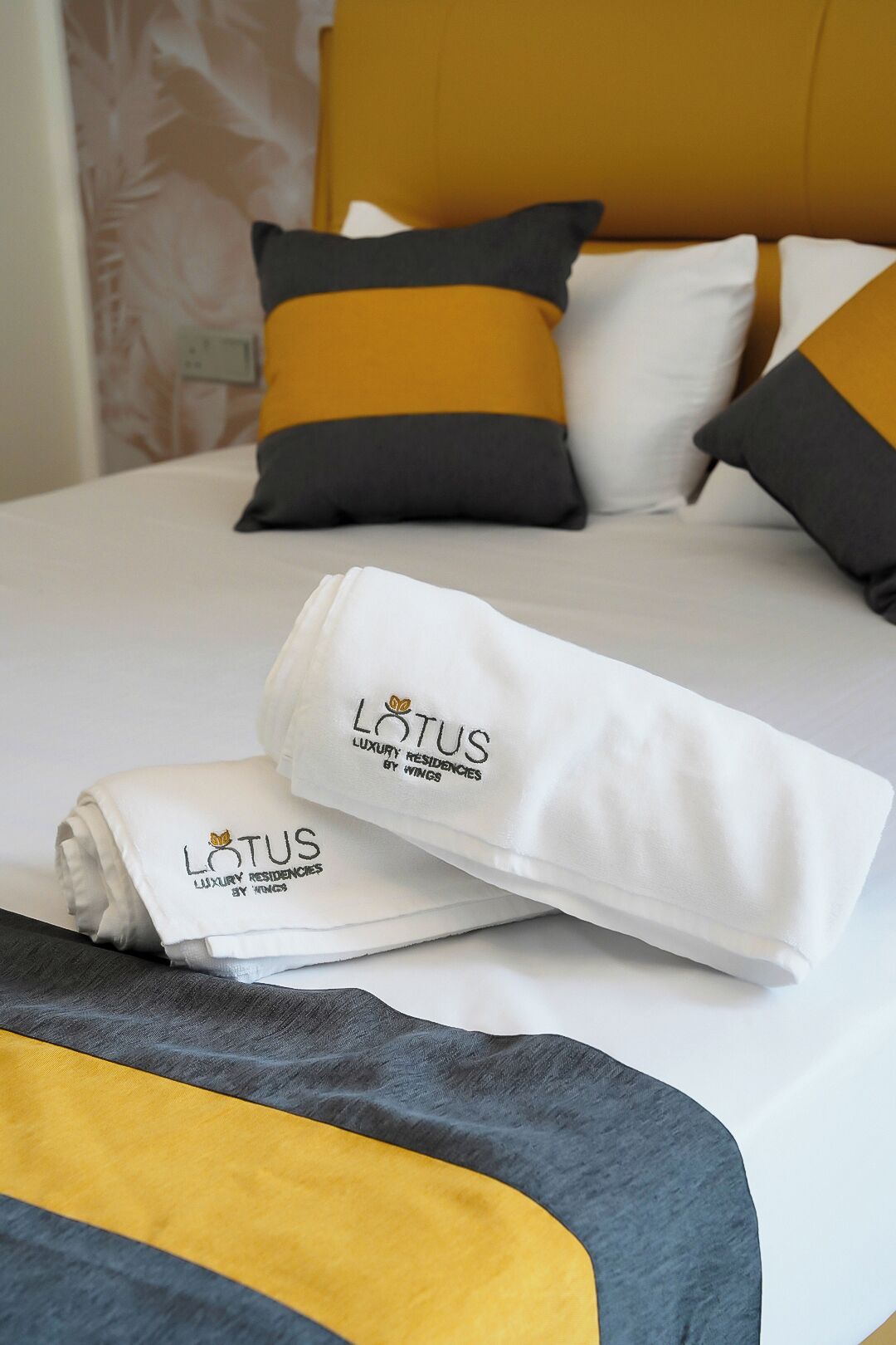 A neatly made bed with a white and yellow bedspread, a folded white towel with the logo 'LOTUS LUXURY RESIDENCIES BY WINGS', and matching pillows.