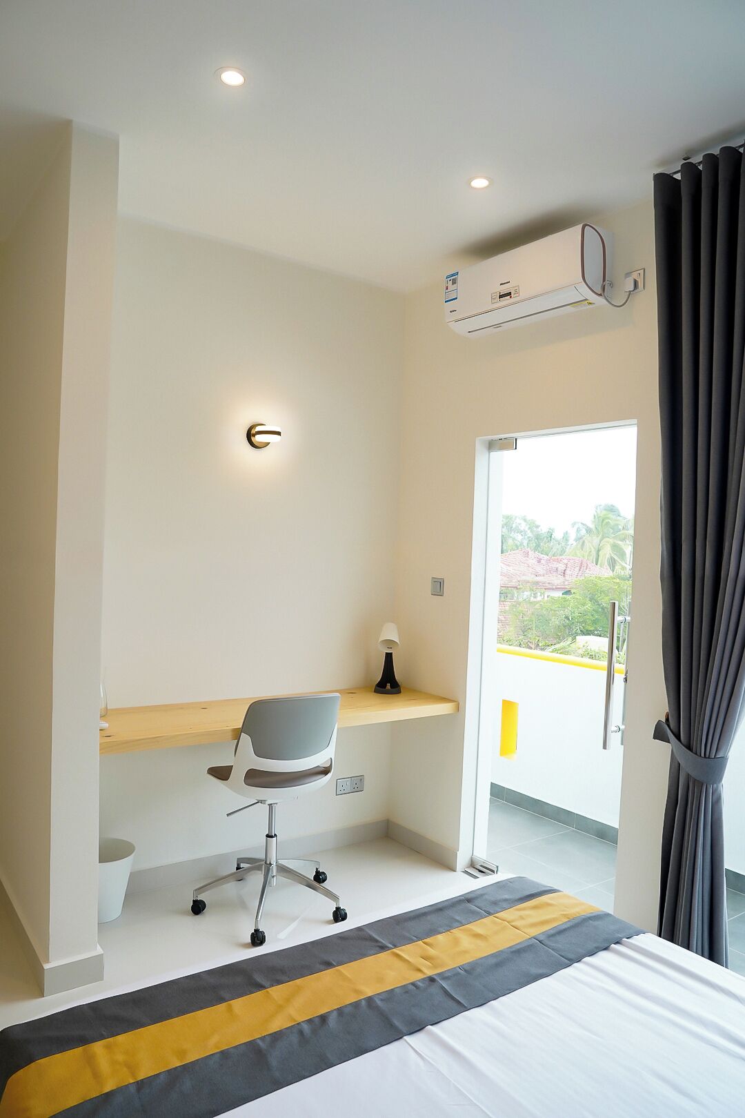 A modern hotel room with a bed, a desk with a chair, an air conditioning unit, and a view of greenery outside.