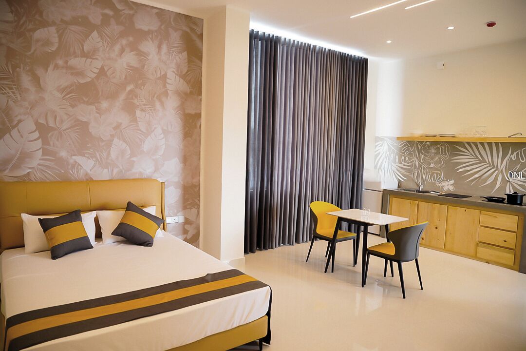 A modern hotel room with a large bed, a small table with chairs, a kitchen area, and a curtain separating the dining area from the bedroom.