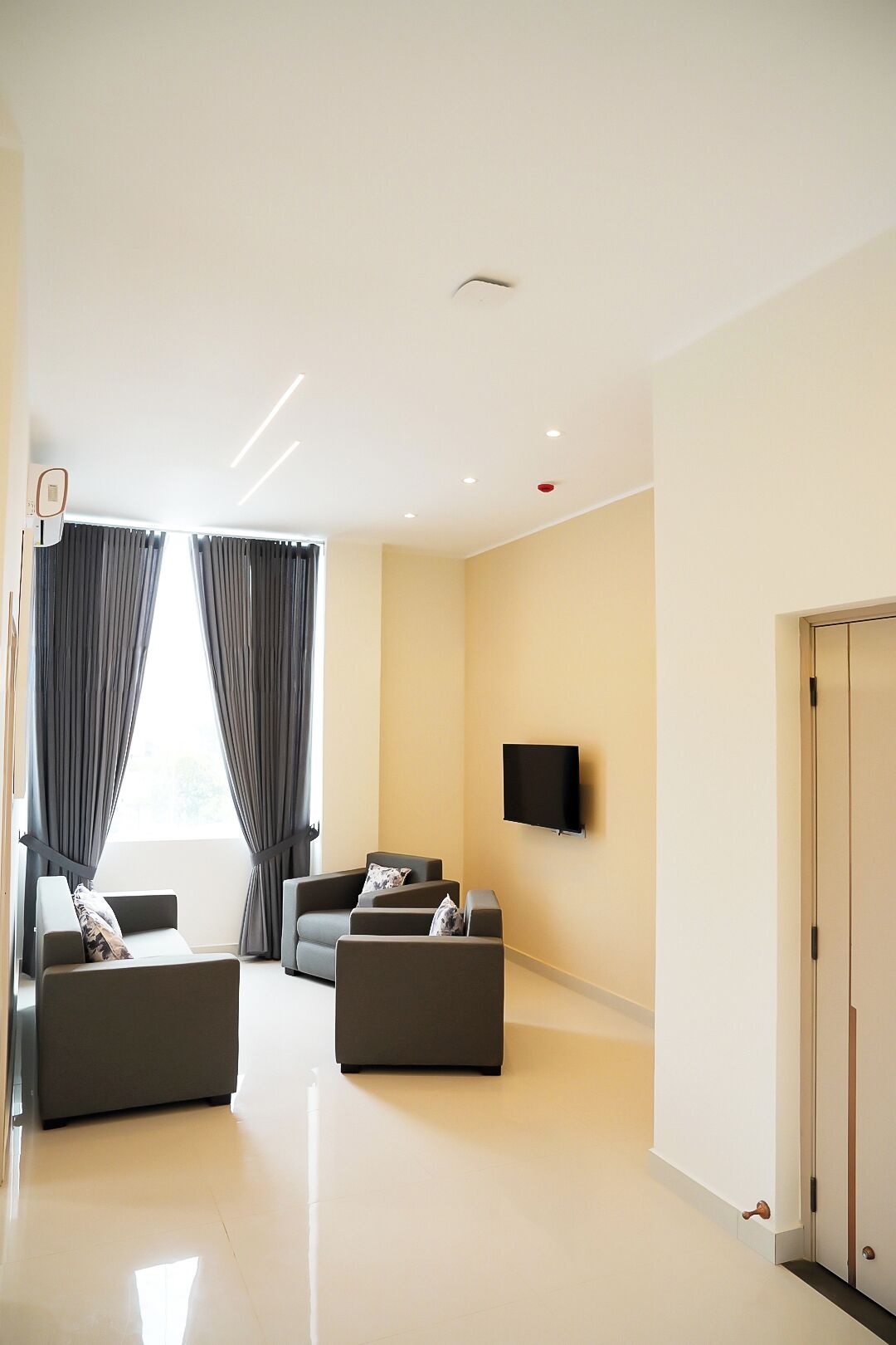 Uma sala de estar moderna com uma televisão de tela plana na parede, dois sofás cinzas com almofadas, uma janela grande com cortinas cinzas e uma porta à direita.