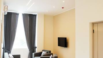 Uma sala de estar moderna com uma televisão de tela plana na parede, dois sofás cinzas com almofadas, uma janela grande com cortinas cinzas e uma porta à direita.