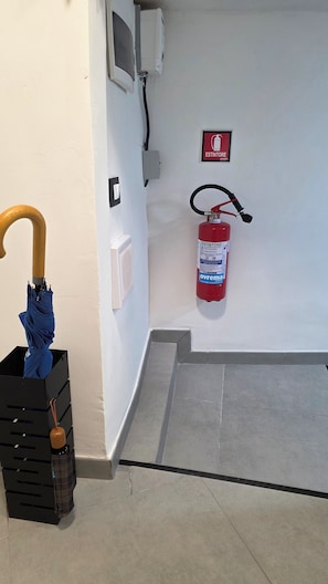 Ein Flur mit an der Wand montiertem Feuerlöscher, einem Holzschirm, der an einem schwarzen Stauraumschrank lehnt, und einer weißen Wand mit Steckdose und kleinem Stromverteiler.