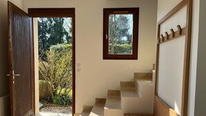 Un interior moderno con una puerta de madera que da a un jardín, una ventana pequeña y una escalera con barandilla de madera.