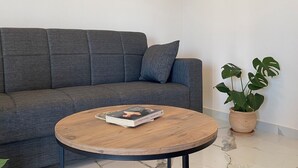 A grey sofa with a single pillow, a round wooden coffee table with a book, and a potted plant with large leaves.