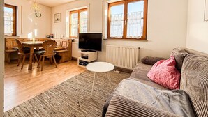 Ein gemütliches Wohnzimmer mit einer Couch, einem Fernseher, einem Essbereich mit Tisch und Stühlen sowie großen Fenstern.