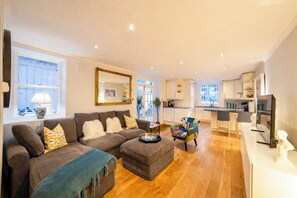 A modern living room with a sofa, ottoman, and a colorful chair. There is a kitchen area with a bar and a dining table. - Beautifully renovated 2-bedroom, 2-bathroom garden apartment in the heart of Kensington & Chelsea, featuring a spacious king en-suite, flexible bedding, bright living areas, and elegant finishes — perfect for families, friends, or business stays. (Greater London)