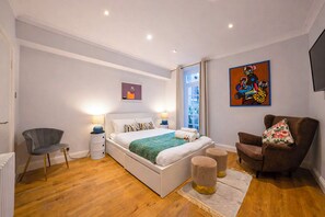 A modern bedroom with a large bed, bedside lamps, a chair, a sofa, and artwork on the walls. - Beautifully renovated 2-bedroom, 2-bathroom garden apartment in the heart of Kensington & Chelsea, featuring a spacious king en-suite, flexible bedding, bright living areas, and elegant finishes — perfect for families, friends, or business stays. (Greater London)