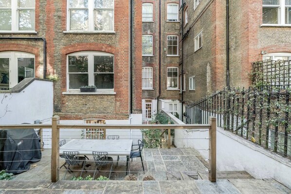 A courtyard with a table and chairs, a brick building, and a metal fence. - Beautifully renovated 2-bedroom, 2-bathroom garden apartment in the heart of Kensington & Chelsea, featuring a spacious king en-suite, flexible bedding, bright living areas, and elegant finishes — perfect for families, friends, or business stays. (Greater London)