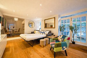 A modern bedroom with a bed, a round mirror, a sofa, a colorful armchair, and a dining area with chairs. - Beautifully renovated 2-bedroom, 2-bathroom garden apartment in the heart of Kensington & Chelsea, featuring a spacious king en-suite, flexible bedding, bright living areas, and elegant finishes — perfect for families, friends, or business stays. (Greater London)