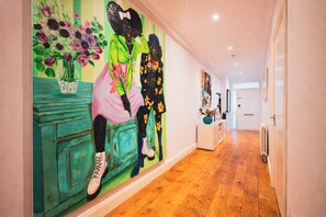 A hallway with a mural of two figures, a white door, and a white console table with a vase. - Beautifully renovated 2-bedroom, 2-bathroom garden apartment in the heart of Kensington & Chelsea, featuring a spacious king en-suite, flexible bedding, bright living areas, and elegant finishes — perfect for families, friends, or business stays. (Greater London)