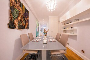 A modern dining area with a long table, chairs, and a decorative wall piece. - Beautifully renovated 2-bedroom, 2-bathroom garden apartment in the heart of Kensington & Chelsea, featuring a spacious king en-suite, flexible bedding, bright living areas, and elegant finishes — perfect for families, friends, or business stays. (Greater London)