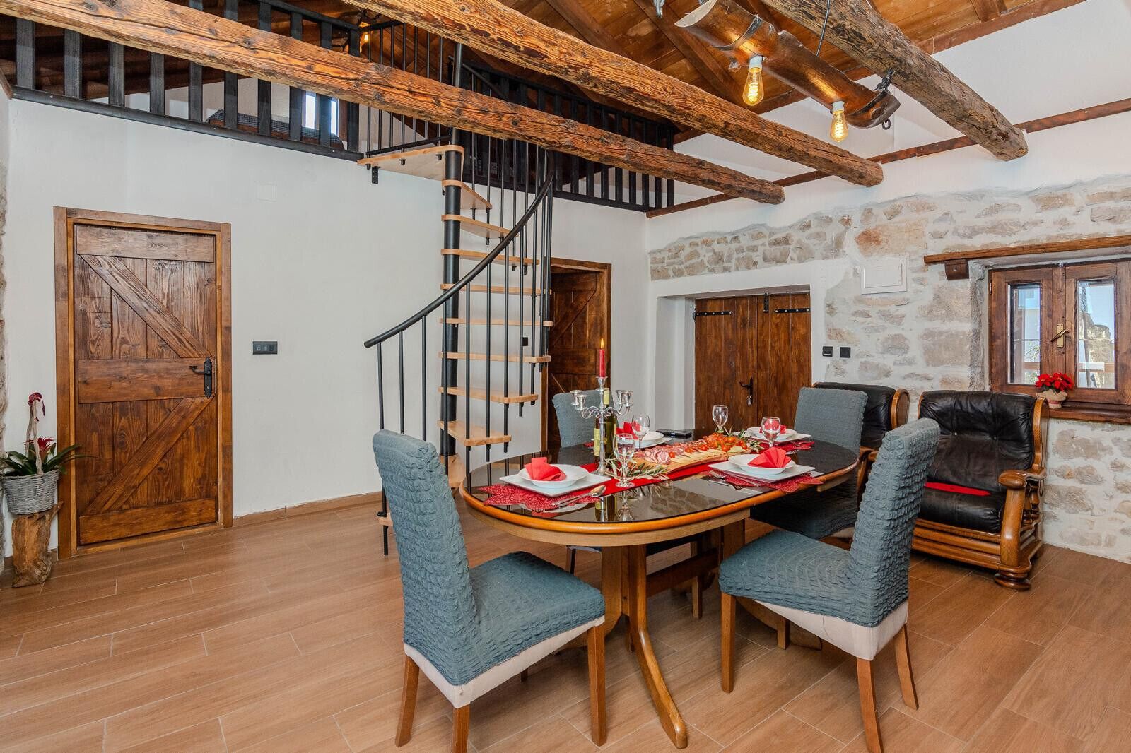 Un comedor con una mesa de madera dispuesta para cuatro personas, rodeada de sillas tapizadas. En el fondo, se ve una escalera en espiral que conduce a un nivel superior.