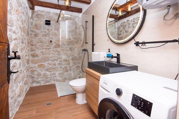 A compact bathroom with a stone wall, a wooden floor, a white toilet, a washing machine, and a sink with a black countertop. - Villa Niko Zadarvillas (Škabrnja)