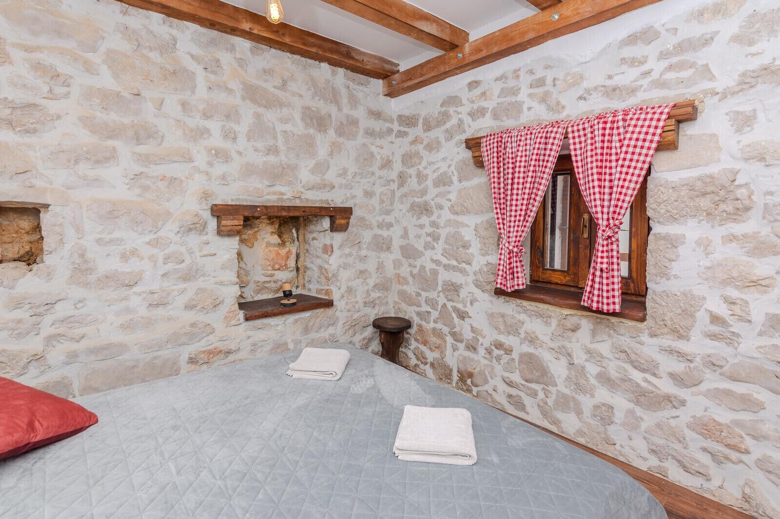 A stone-walled room with a bed, a small wooden shelf, and a window with red and white checkered curtains.