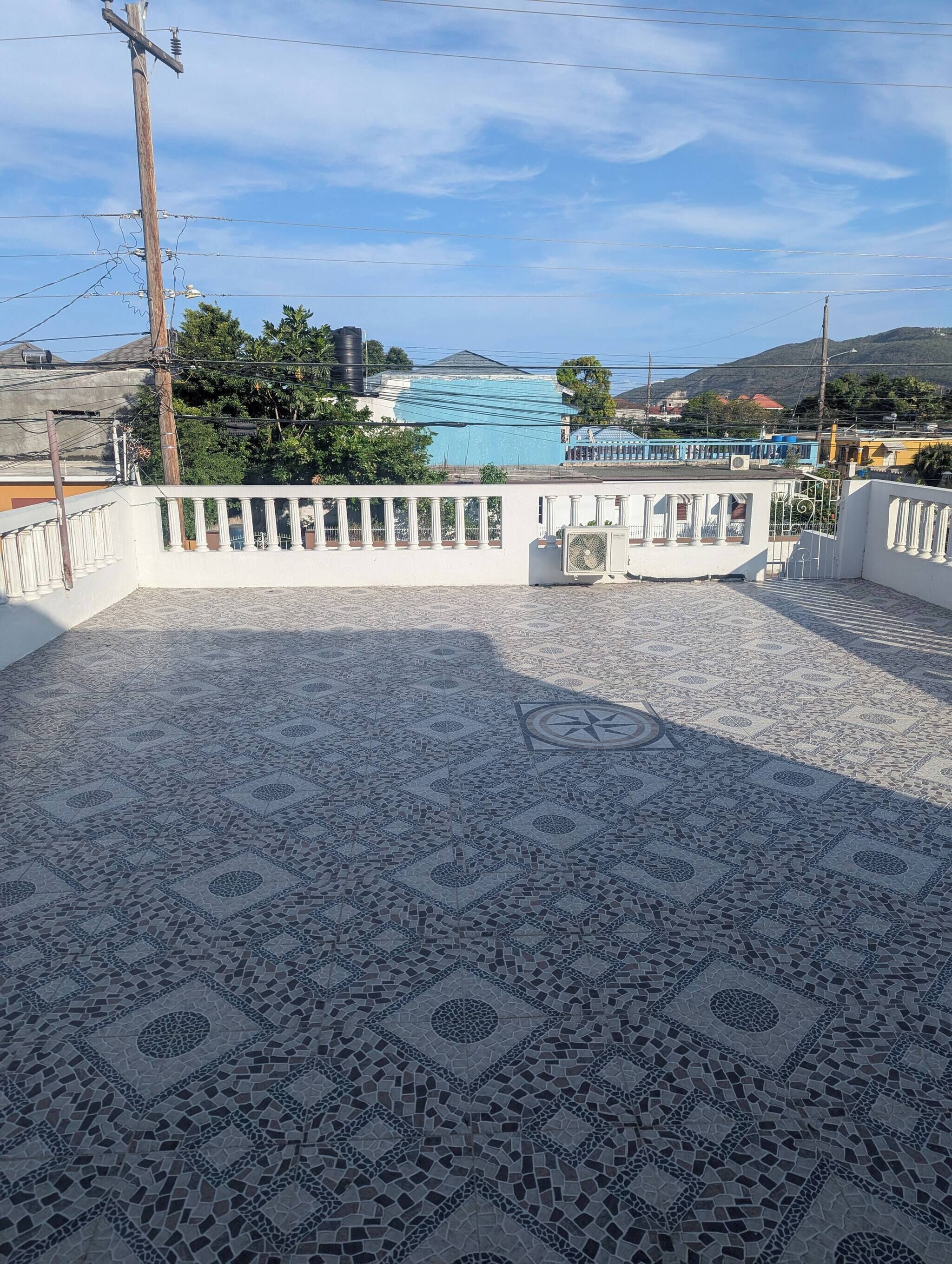 Una terrazza piastrellata con pavimento decorato, ringhiera bianca e vista su un edificio con tetto blu e uno specchio d'acqua sullo sfondo.