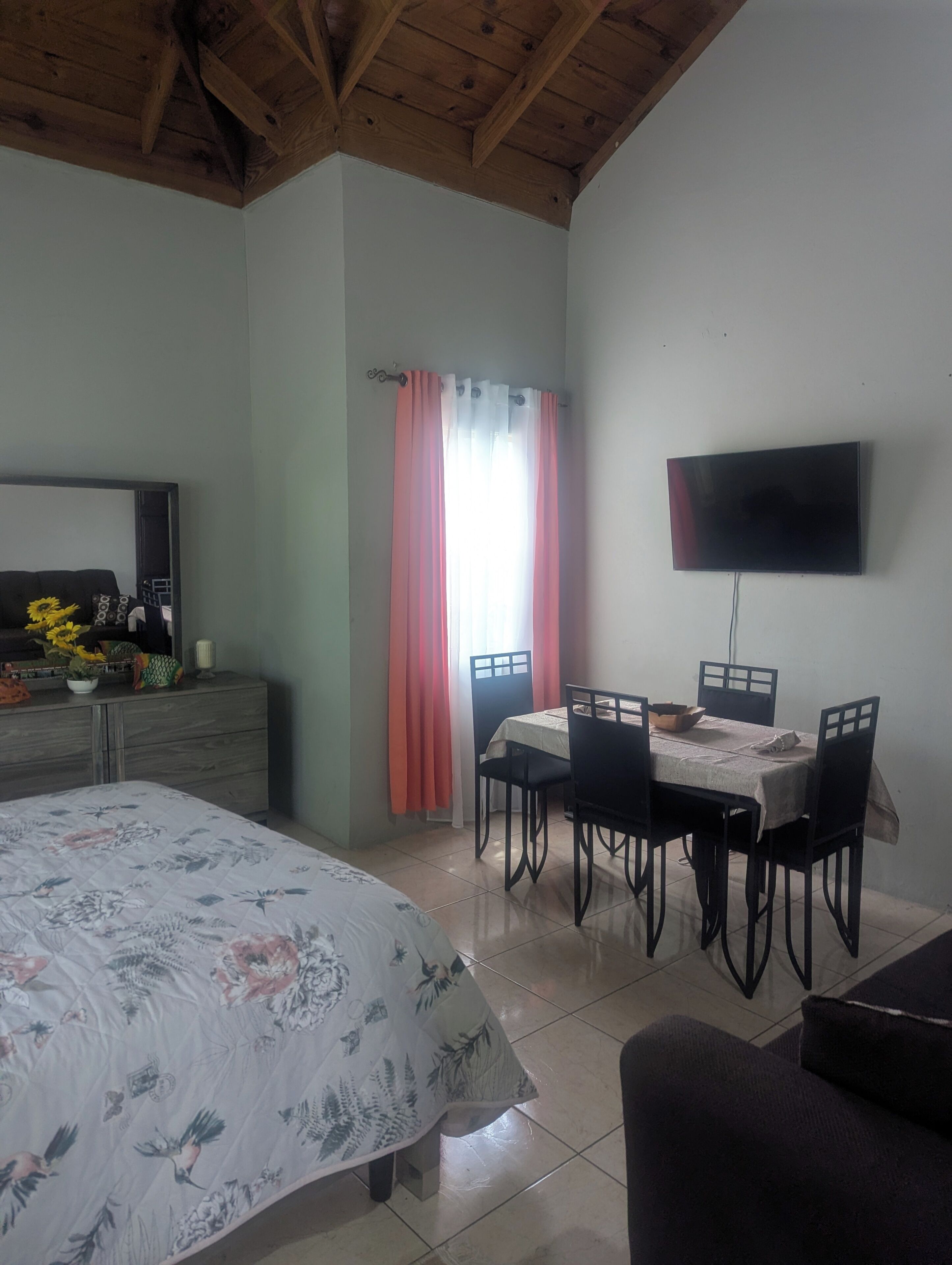 Un dormitorio con cama, televisión montada en la pared, mesa de comedor con sillas y ventana con cortinas.
