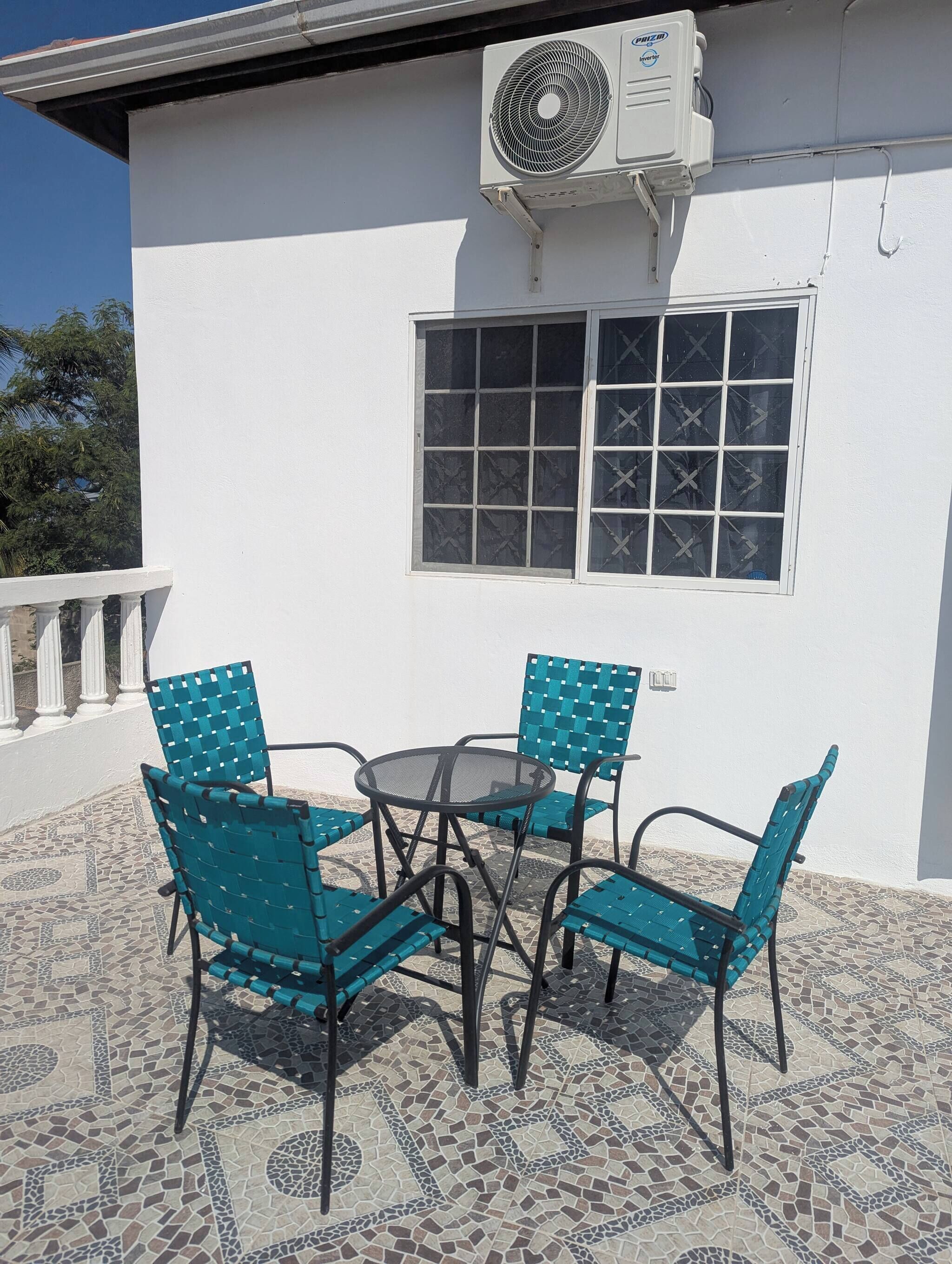 Une terrasse extérieure avec une table et quatre chaises, une fenêtre avec des barreaux en métal et un appareil de climatisation fixé au mur.