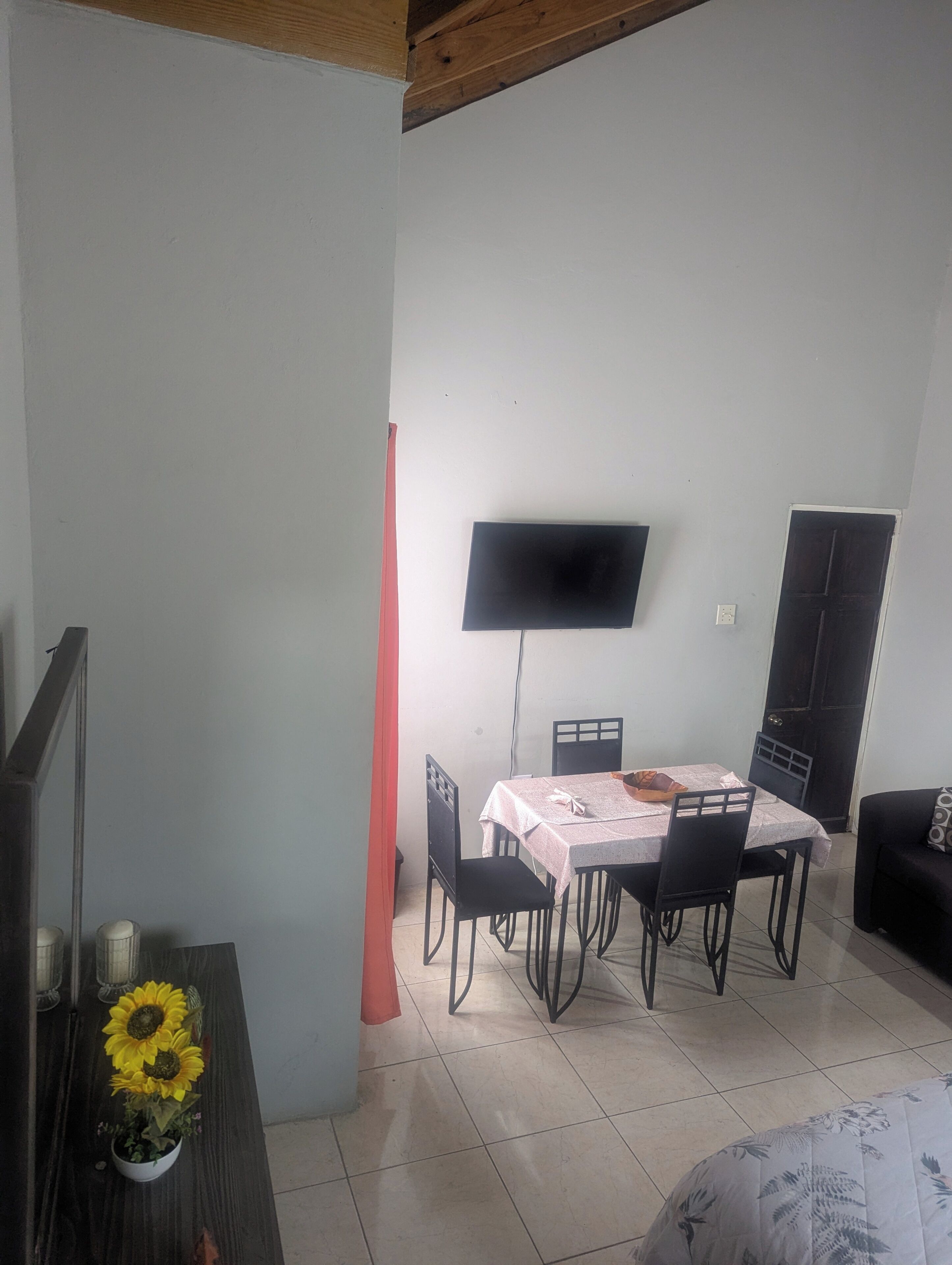 A dining area with a table set for four, a flat-screen TV mounted on the wall, and a dark-colored sofa in the corner.