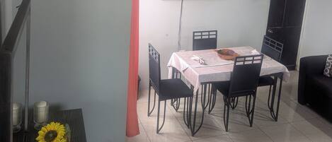 A dining area with a table set for four, a flat-screen TV mounted on the wall, and a dark-colored sofa in the corner.