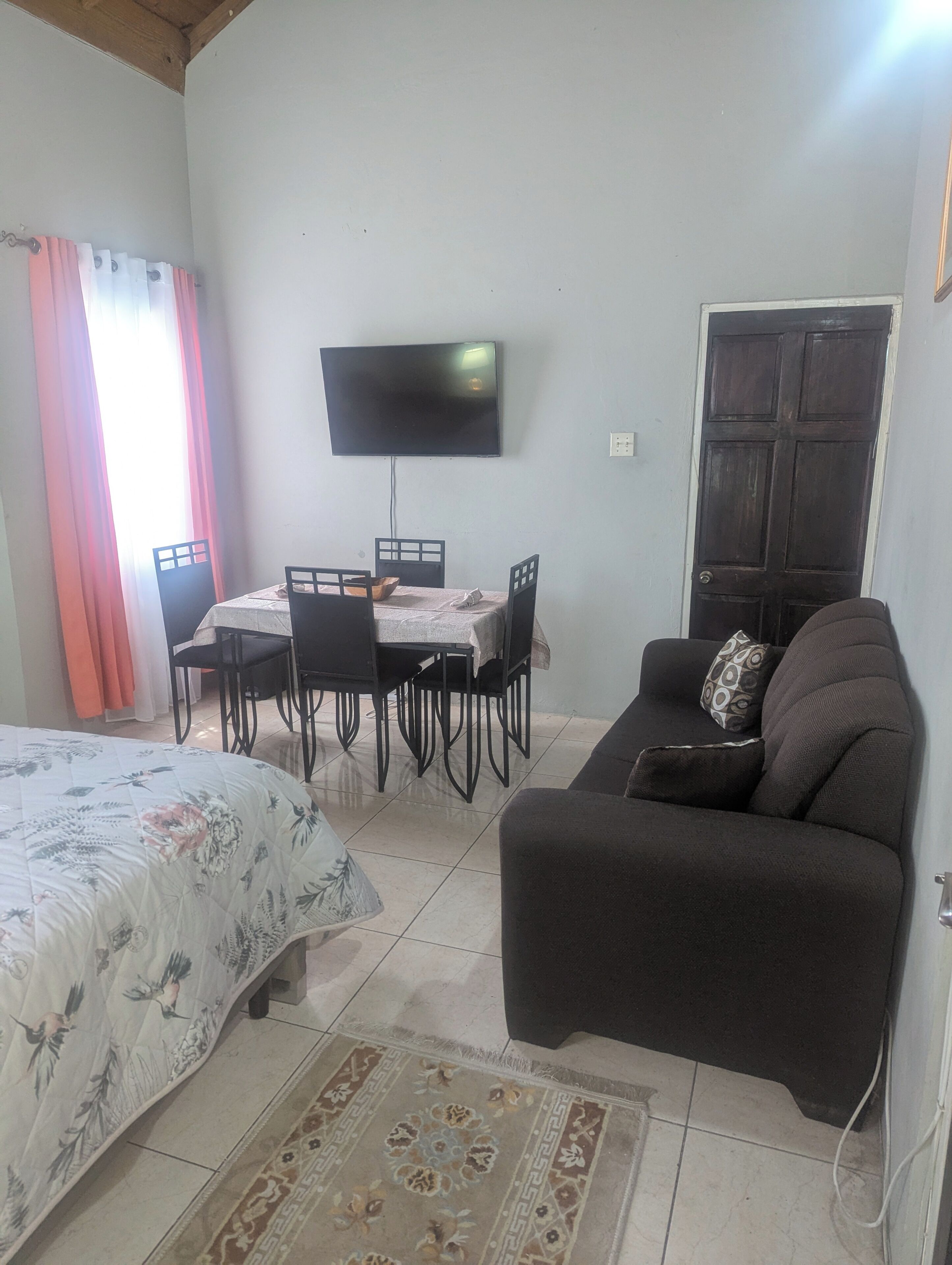 A room with a bed, a sofa, a dining table with chairs, a TV mounted on the wall, and a door.