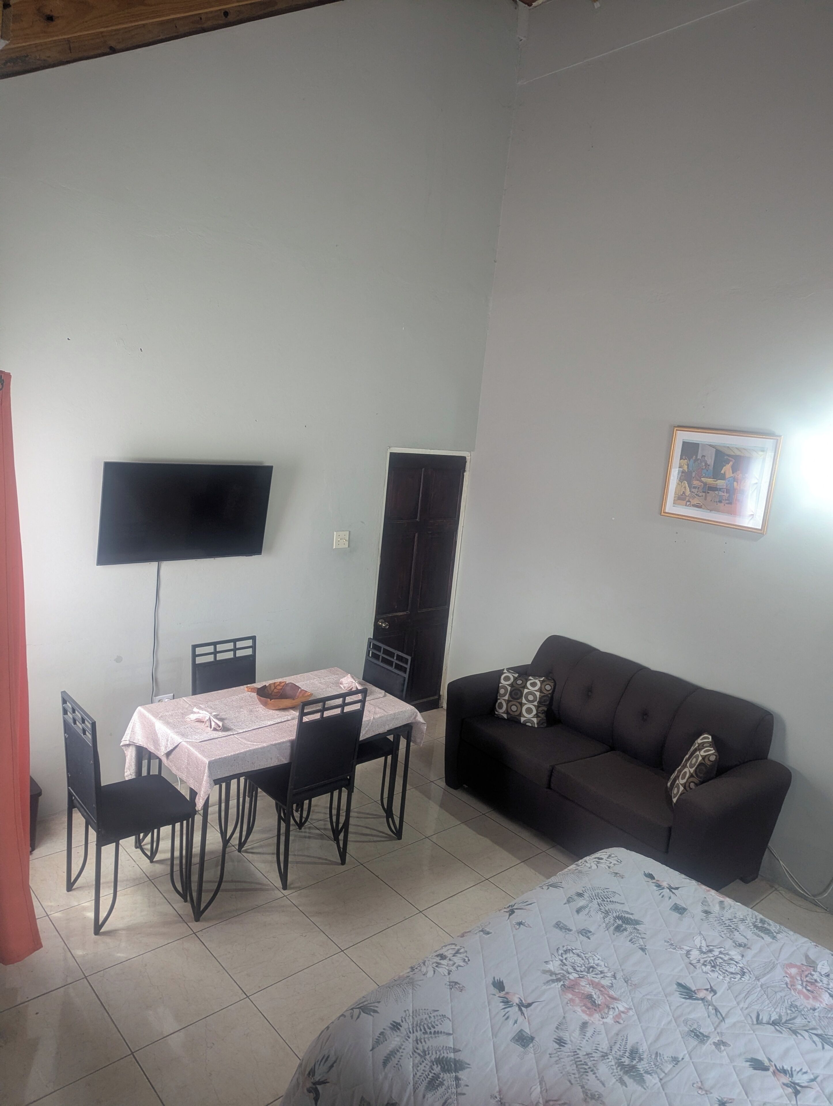 A room with a dining table, chairs, a black sofa, a bed with a floral duvet, a TV mounted on the wall, and a door.