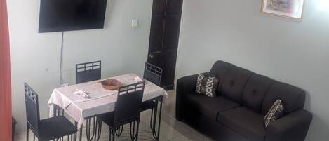 A room with a dining table, chairs, a black sofa, a bed with a floral duvet, a TV mounted on the wall, and a door.