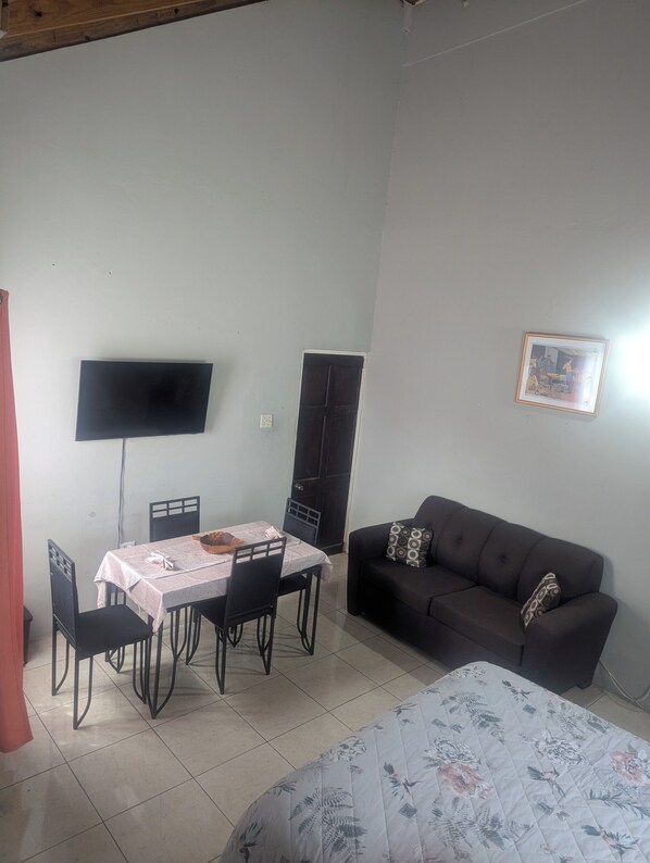 A room with a dining table, chairs, a black sofa, a bed with a floral duvet, a TV mounted on the wall, and a door. - 2 Bedroom Upstairs Large Flat in Portmore Pines (Portmore)