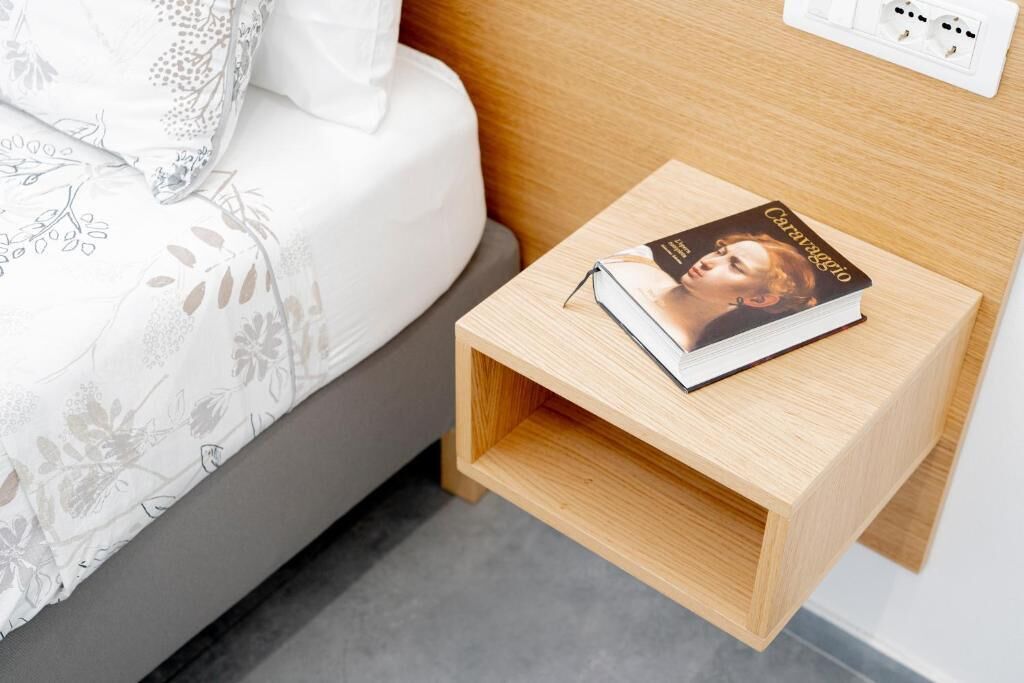 A neatly made bed with a floral-patterned bedspread and a pillow. A small wooden nightstand with a book titled "Caravaggio" is placed beside the bed.
