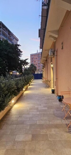 A corridor with a tiled floor, bordered by a low wall with plants, leading to a building with balconies. - Eos Elegant Apartment & Suites (Syracuse)