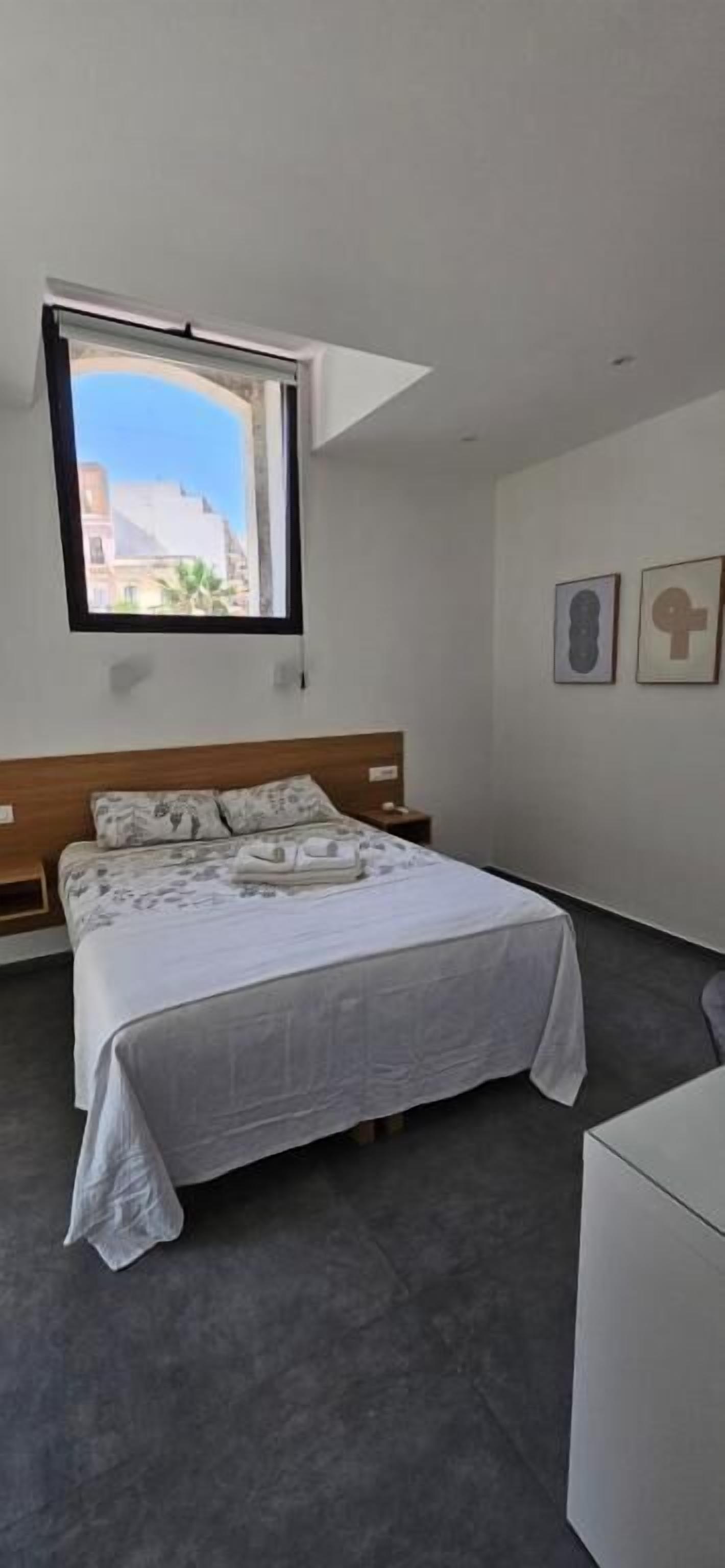 Un dormitorio con cama, ventana, mesita de noche y un pequeño armario blanco.