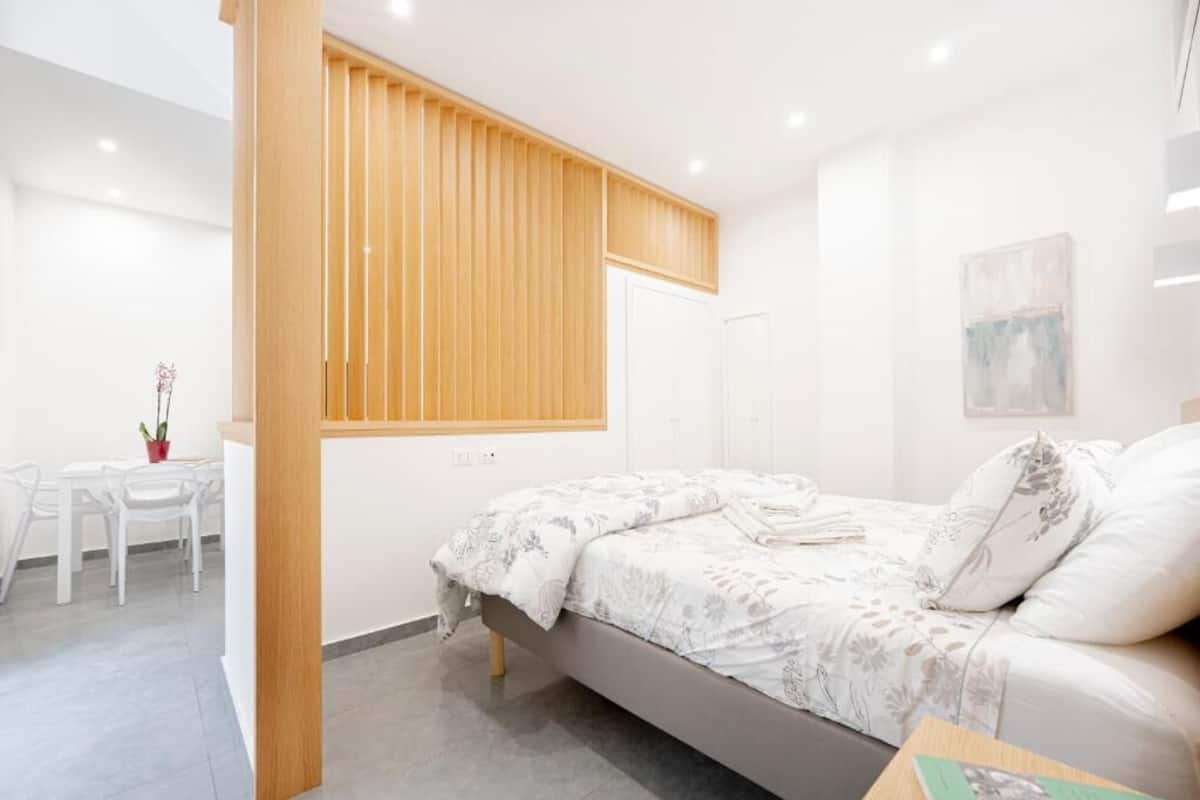 Een moderne slaapkamer met een bed, een raam met houten lamellen, een wit bureau en een stoel.