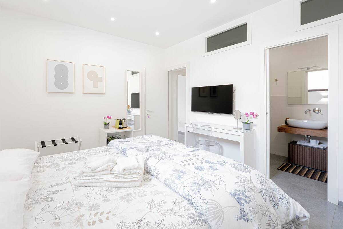 Een moderne slaapkamer met een groot bed, een televisie, een badkamer met een wastafel en spiegel, en kunst aan de muur.