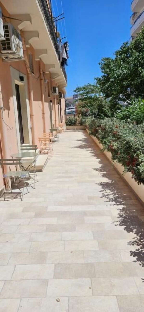 A corridor with a tiled floor, outdoor seating, and a row of buildings with balconies. - Eos Elegant Apartment & Suites (Syracuse)