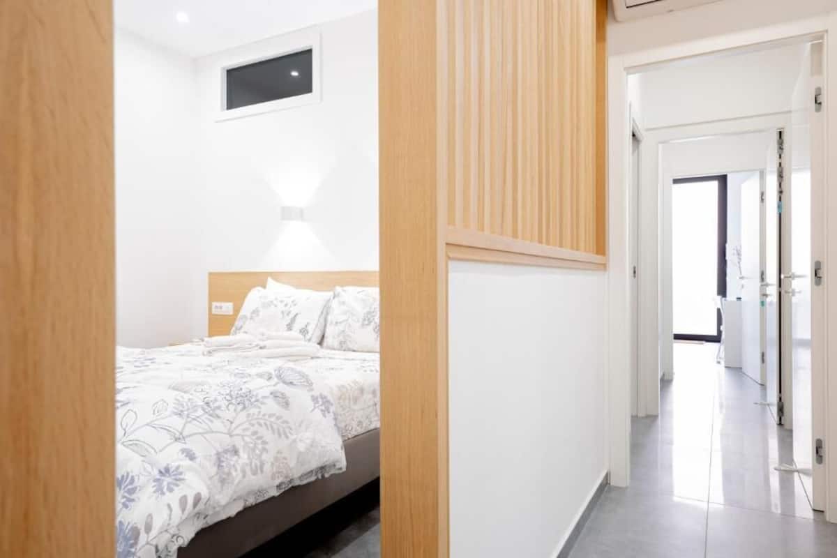Een moderne slaapkamer met een bed, een nachtkastje en een bijpassend tafeltje. Er is een raam met jaloezieën, een wandlamp en een deur naar een andere kamer.